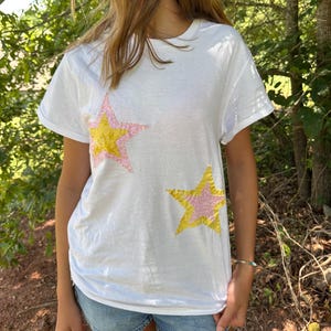 May include: White t-shirt with two star-shaped appliqués. Each star features a yellow center and a pink glitter outline. The shirt has short sleeves and a crew neck, suitable for casual wear.