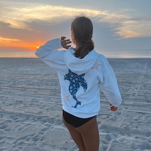 May include: White hooded sweatshirt with a blue dolphin design on the back and pink star accents. The person is standing on a sandy beach, facing the sunset over the ocean. The sky is a mix of orange, yellow, and blue.