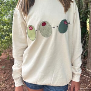 May include: Cream-colored sweatshirt with three olive appliqués. Each olive is a different shade of green, with a red or checkered accent. The olives are connected by a thin black line. The sweatshirt has long sleeves and a crew neck.