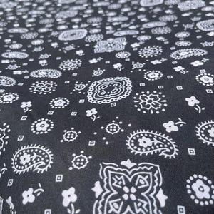 May include: Black fabric with a white paisley pattern. The design includes various floral and geometric shapes, such as paisley, flowers, and small squares, creating a repeating pattern across the material.