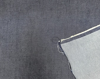 May include: A piece of dark indigo blue denim fabric with a visible twill weave pattern. The fabric is folded over to reveal a lighter, un-dyed white interior side with a distinct diagonal texture and a raw, frayed edge along the fold line.