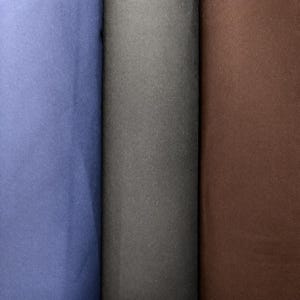 Puede incluir: Tres rollos de tela en tonos azul, gris y marrón. La tela parece ser de color liso, con una textura suave. Los rollos están bien enrollados y los bordes son visibles.