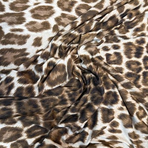 May include: Close-up of a fabric with a leopard print design. The material features a white base with brown and dark brown spots. The fabric is folded, displaying the texture and pattern of the material.