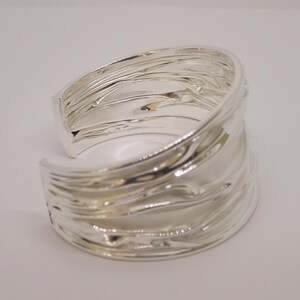 May include: A wide, silver-colored cuff bracelet with a textured, rippled design. The open-ended bracelet has a smooth, polished interior and a wave-like exterior, reflecting light. The jewelry piece is set against a plain white background.