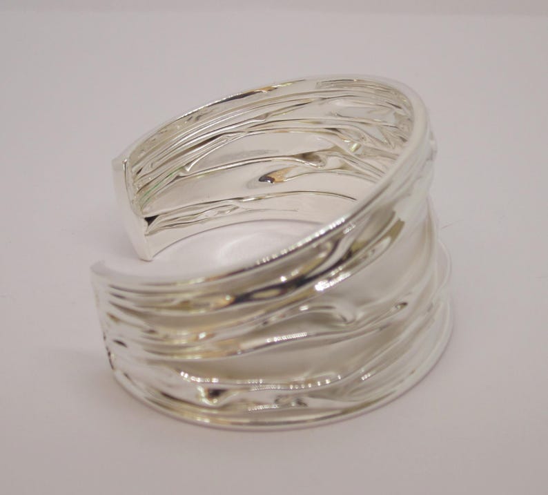 May include: A wide, silver-colored cuff bracelet with a textured, rippled design. The open-ended bracelet has a smooth, polished interior and a wave-like exterior, reflecting light. The jewelry piece is set against a plain white background.