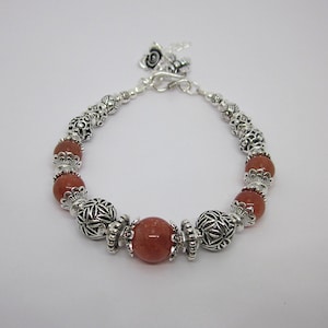 May include: A beaded bracelet featuring silver-colored metal beads and reddish-brown gemstone beads. The bracelet has a decorative clasp and a small charm. The beads are round and vary in size, creating a unique jewelry piece.