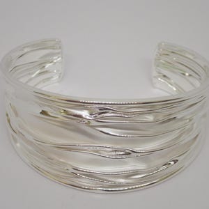 May include: A wide, open silver cuff bracelet with a textured, wavy design. The bracelet has a polished, reflective surface and is made of a shiny metal. The jewelry piece is a statement accessory.