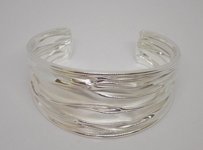 May include: A wide, open silver cuff bracelet with a textured, wavy design. The bracelet has a polished, reflective surface and is made of a shiny metal. The jewelry piece is a statement accessory.