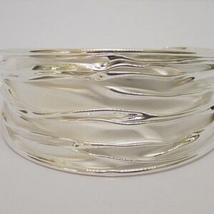 May include: A wide, silver-colored cuff bracelet with a rippled, wave-like design. The bracelet has a smooth, reflective surface, and the light catches the curves, creating highlights and shadows. The overall design is modern and elegant.