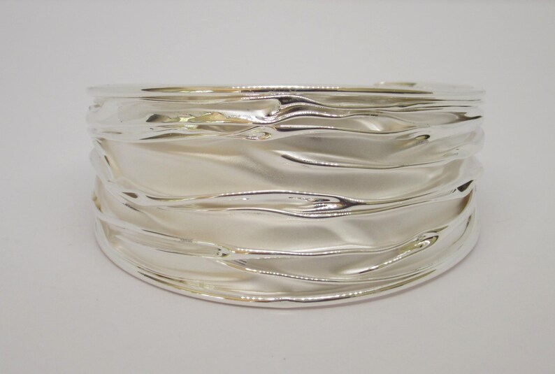 May include: A wide, silver-colored cuff bracelet with a rippled, wave-like design. The bracelet has a smooth, reflective surface, and the light catches the curves, creating highlights and shadows. The overall design is modern and elegant.