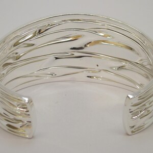 May include: A silver-colored cuff bracelet with a textured, wave-like design. The open-ended bracelet has a smooth interior and a series of parallel, curved lines on the exterior. The metal reflects light, creating highlights and shadows.