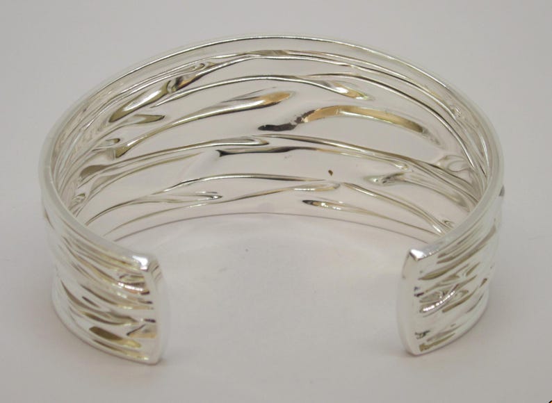 May include: A silver-colored cuff bracelet with a textured, wave-like design. The open-ended bracelet has a smooth interior and a series of parallel, curved lines on the exterior. The metal reflects light, creating highlights and shadows.