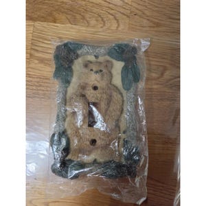 May include: A rectangular light switch wall plate featuring a sculpted brown bear in the center, surrounded by a border of green pine needles and brown pinecones. The plate has two circular screw holes and a central rectangular toggle switch cutout.