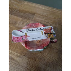 May include: A white ceramic spoon with a butterfly motif on the handle, attached to a rectangular card labeled Garden Collection By CornerRuby, resting on a pink and red floral patterned ceramic saucer wrapped in clear plastic packaging.