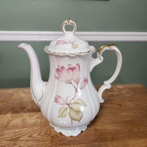 May include: White porcelain coffee pot with a lid, featuring a pink floral design. The pot has a curved spout, a gold-colored handle, and a decorative gold trim. The lid has a small, ornate handle.