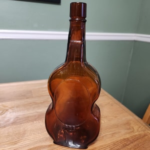 May include: A brown glass bottle shaped like a violin. The bottle has a long neck and a rounded body with a detailed design. The glass has a translucent quality, allowing light to pass through.