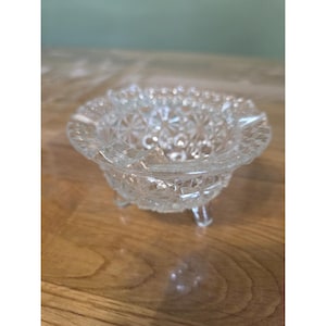 May include: A clear glass ashtray featuring a pressed starburst and diamond pattern across its exterior surface. The circular bowl is supported by three small, integrated glass feet and has a scalloped rim with notches for resting items.