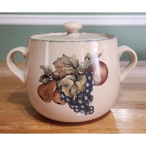 May include: Cream-colored ceramic casserole dish with lid and two handles. The dish features a fruit design with grapes, pears, and an apple. The lid has a small knob. The rim has a blue trim.