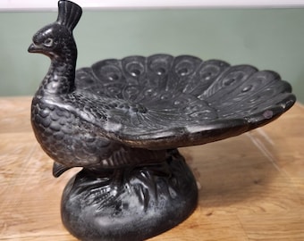 Puede incluir: Un plato decorativo en forma de pavo real gris oscuro. El pavo real tiene un diseño de plumas detallado y una base pequeña y redonda. El plato está hecho de un material pesado y se encuentra sobre una superficie de madera.