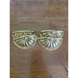 May include: Four gold-colored, half-circle drawer pulls. Each pull has a fan-like design with vertical lines radiating from the center. The pulls are made of a shiny, metallic material and are arranged on a wooden surface.