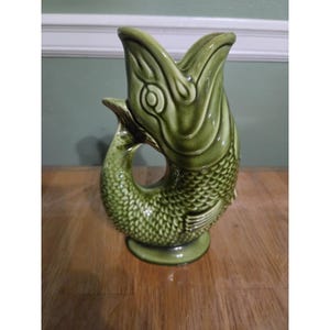 May include: A glossy olive green ceramic pitcher shaped like a fish with its mouth open at the top. The body features detailed scale textures, a curved tail forming a handle, and a circular base. The glaze has darker green shading in the crevices.