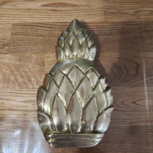 May include: A gold-colored, pineapple-shaped decorative object. The pineapple has a textured surface with a series of diamond-shaped segments. The object is made of metal and has a shiny finish. The pineapple is a symbol of hospitality.