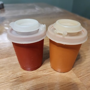 May include: Two small, cylindrical containers with white lids. One is a deep red color, and the other is orange. The lids have a hinged design for easy opening and closing. These containers are likely for spices or condiments.