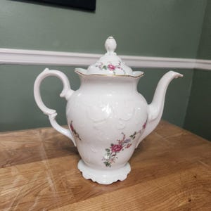 May include: White porcelain teapot with a lid, featuring embossed floral patterns and delicate pink and purple rose accents. The teapot has a gold trim and a curved handle, perfect for serving tea.