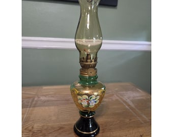 Puede incluir: Una lámpara de aceite con una chimenea de cristal verde translúcido, un mecanismo de quemador de latón y una base de cristal bulbosa. La base es de color ámbar con motivos florales pintados a mano en blanco, azul y verde sobre un pedestal negro.