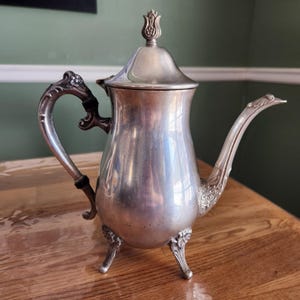 May include: A silver-colored antique coffee pot with a curved spout and ornate handle. The pot has a lid with a tulip-shaped finial and rests on three decorative feet. The handle has a black accent.