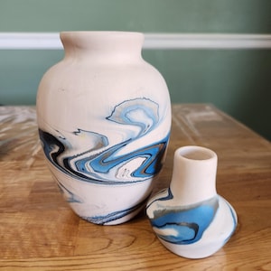 May include: Two ceramic vases. The larger vase is a light pink with blue, black, and gray swirled patterns. The smaller vase has a similar design. Both vases have a smooth, matte finish and are displayed on a wooden surface.