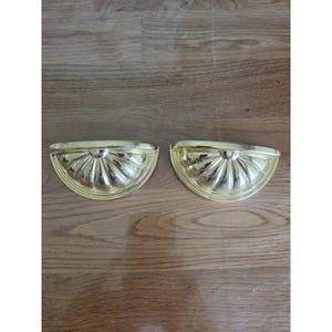 May include: Two gold-colored, half-circle drawer pulls with a fluted design. The pulls have a shiny finish and are set against a wooden surface. These decorative hardware pieces are likely for furniture.