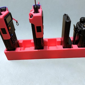 May include: A red plastic organizer holding four black two-way radios. The radios are of different sizes and shapes. The organizer has four slots to hold the radios upright. The radios have antennas and some have attached lanyards.