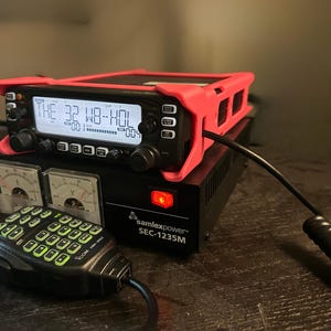 May include: A black and red radio transceiver with the text "THE 32 WB-HOL" displayed on the screen. The radio is mounted on a black power supply with a red power button and two analog meters. A black microphone is connected.