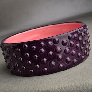May include: A dark purple ceramic bowl with a textured exterior of small, rounded bumps. The interior is a bright pink color. The oval-shaped bowl sits on a gray surface. This decorative bowl is suitable for home decor.