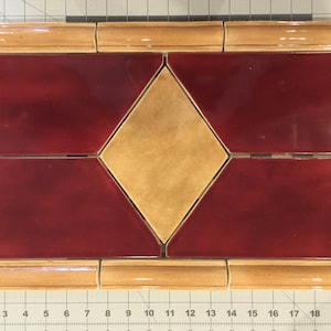 May include: A tile design featuring a diamond shape in the center with a light brown glaze. The diamond is surrounded by four red tiles with a darker brown glaze. The entire design is framed by a border of light brown tiles.