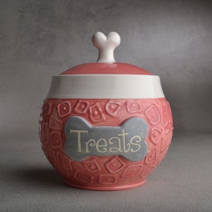 Op de afbeelding: Roze keramische hondenkoekjespot met een wit, botvormig deksel en een grijs bot met het woord "Treats" erop.