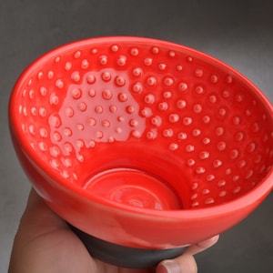 Puede incluir: Un cuenco de cerámica rojo brillante con una superficie texturizada y con hoyuelos. El cuenco tiene una base negra.