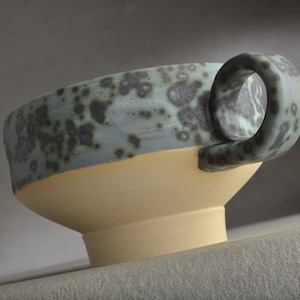 Puede incluir: Una taza de cerámica con un esmalte verde moteado y un esmalte amarillo en la parte inferior. La taza tiene un asa.