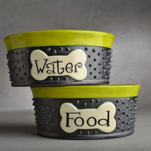 May include: Two ceramic pet bowls, one green and one black, with a bone-shaped label on each. The top bowl says "Water" and the bottom bowl says "Food".