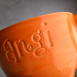 Puede incluir: Taza de cerámica naranja con la palabra "angi" en relieve en el lateral.