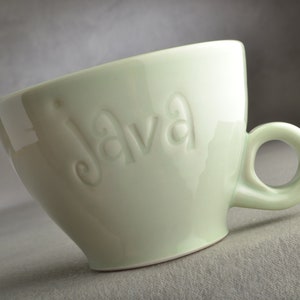 Könnte beinhalten: Eine hellgrüne Keramiktasse mit abgerundeter Form und einem Henkel. Das Wort "Java" ist auf der Vorderseite der Tasse geprägt.