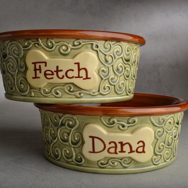 May include: Two ceramic dog bowls with green and brown glaze. The top bowl has the name "Fetch" on it and the bottom bowl has the name "Dana" on it.