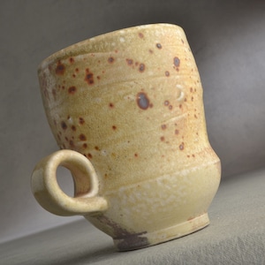 May include: A handmade ceramic mug with a speckled yellow glaze and a brown handle. The mug has a unique, slightly asymmetrical shape.
