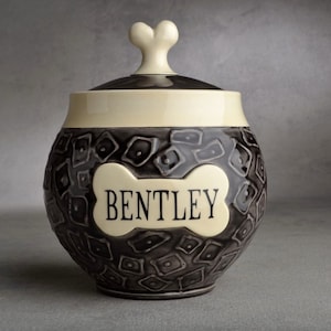 Puede incluir: Una urna de cerámica para mascotas con una tapa en forma de hueso y una placa en forma de hueso que dice "BENTLEY". La urna está decorada con un patrón geométrico en blanco y negro.