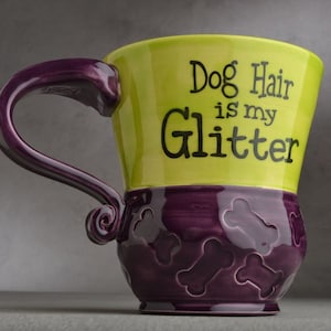 Puede incluir: Una taza de cerámica verde y morada con asa. La taza tiene el texto "Dog Hair is my Glitter" en la parte delantera. La parte inferior de la taza está decorada con formas de huesos.