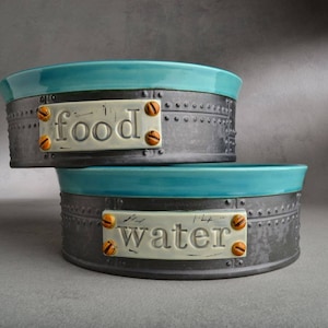 May include: Two ceramic pet bowls with teal blue interiors and a dark gray exterior. The bowls have metal accents and labels that read "food" and "water".