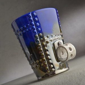 May include: A blue ceramic mug with a textured, metallic finish and a white ceramic handle shaped like a wheel. The mug has a unique design with small, raised dots.