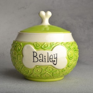 May include: Green ceramic pet food container with a white bone-shaped design and the name "Bailey" written in black. The container has a lid with a bone-shaped knob.