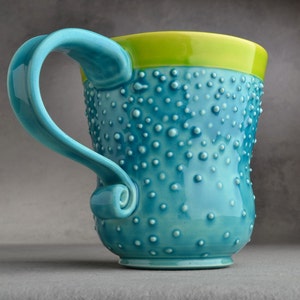 Peut inclure: Une tasse en céramique bleu turquoise avec un bord vert et une surface texturée. La tasse a une poignée incurvée unique.